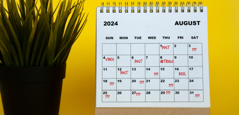 un calendario editoriale social su calendario da tavolo e accanto una pianta da ufficio. Sul calendario nelle diverse date sono segnati i vari contenuti da condividere e in molte date dei punti di domanda